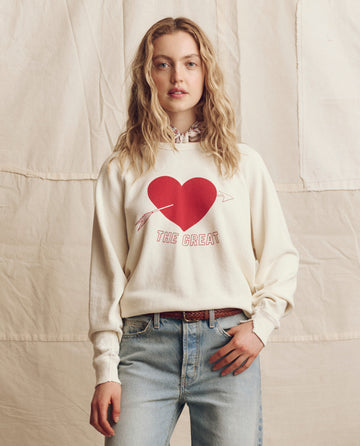 A woman stands before a beige backdrop in Los Angeles, wearing THE GREAT. PS26 The College Sweatshirt in Washed White with a red heart graphic, paired with light blue jeans.