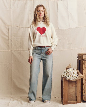 A woman indoors wears THE GREAT. PS26's The College Sweatshirt, Graphic—Washed White with Red Heart, loose blue jeans, and white sneakers beside wooden crates and white flowers for a relaxed Los Angeles vibe.