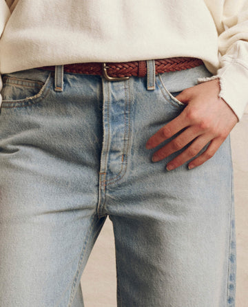 A person wears a cream sweatshirt tucked into THE GREAT. F25 D1 The Slouch Wide Leg Jean. -- Tidal Wash, styled with a brown braided belt and one hand in their pocket.