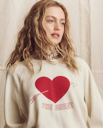 A woman with wavy blonde hair wears THE GREAT. PS26 College Sweatshirt in Washed White, featuring a red heart graphic and "THE GREAT" text, standing before a neutral fabric backdrop inspired by Los Angeles style.