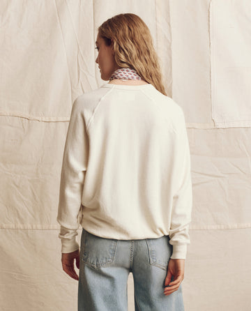 A person with long hair wears THE GREAT. PS26 The College Sweatshirt in washed white with a red heart graphic and blue jeans, standing with their back to the camera before a beige fabric backdrop.