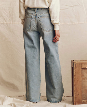 A person stands with their back to the camera wearing THE GREAT. F25 D1 The Slouch Wide Leg Jean in Tidal Wash and a long-sleeve cream top, next to a wooden crate on a neutral fabric backdrop.
