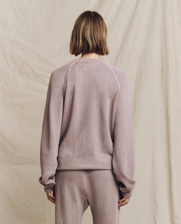 Facing away, the individual is dressed in THE GREAT. HOL 24 KNITS' The Slouch Sweatshirt in Solid Quartz and matching pants, capturing the essence of casual wear against a neutral fabric backdrop.