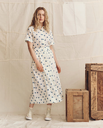 A woman wears THE GREAT. PS26's The Messenger Top in cream with blue polka dots and white sneakers, standing beside two wooden crates against a neutral fabric backdrop.