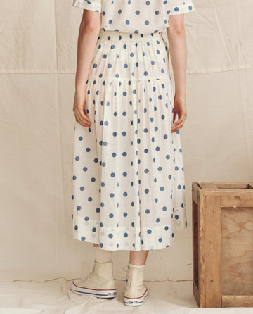 A person wearing THE GREAT. PS26’s The Highland Skirt in cream with blue polka dots stands by a wooden crate, facing away; styled with white socks and sneakers.