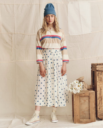 Indoors, a woman wears a blue beanie, white polka dot skirt, white sneakers, and THE GREAT. PS26 The Shrunken Henley. Novelty -- Study Hall Stripe shirt. Wooden crates and a bouquet of flowers sit nearby.