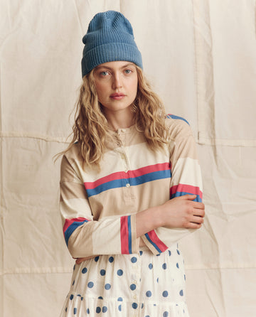 A woman with wavy blonde hair wears THE GREAT. PS26 The Shrunken Henley, Novelty -- Study Hall Stripe, paired with a blue knit beanie and a white skirt with blue polka dots, standing before a light fabric backdrop.