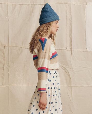 A person in profile stands before a neutral fabric backdrop, wearing THE GREAT. PS26 The Shrunken Henley Novelty -- Study Hall Stripe, paired with a blue knit beanie and a white skirt with blue polka dots.