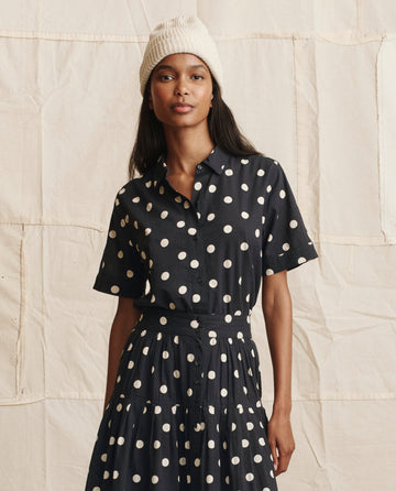 A woman wearing THE GREAT. PS26 The Messenger Top in navy with cream polka dots stands in front of a beige fabric background.