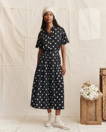 A woman wears THE GREAT. PS26 The Messenger Top in navy with cream polka dots, paired with a white beanie and high-top sneakers, standing before a neutral fabric backdrop with crates and white flowers.