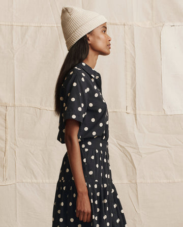 A woman wears THE GREAT. PS26 The Messenger Top in navy with cream polka dots and a cream knit hat, standing in profile against a beige fabric backdrop.