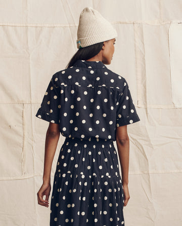 Someone wearing THE GREAT. PS26's The Messenger Top in navy with cream polka dots stands facing away from the camera against a beige fabric backdrop, paired with a cream knit beanie.