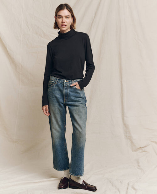 A person wearing The Turtleneck in Almost Black, paired with blue jeans, stands confidently against a plain backdrop, featuring timeless fashion by THE GREAT. HOL 24 TURTLENECKS.