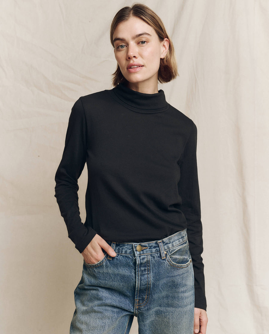 A person wearing The Turtleneck in Almost Black by THE GREAT. HOL 24 TURTLENECKS and blue jeans stands in front of a beige backdrop, embodying effortless fashion.