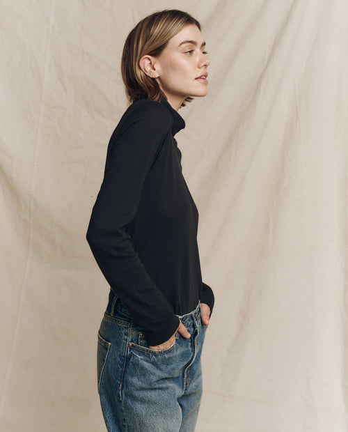 A model wearing The Turtleneck in Almost Black from THE GREAT. HOL 24 TURTLENECKS pairs it with jeans and stands sideways against a beige fabric backdrop, embodying timeless fashion with minimalist elegance.