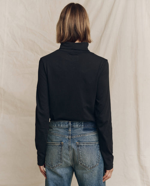 A fashionable person with shoulder-length hair dons *The Turtleneck. -- Almost Black* by THE GREAT. HOL 24 TURTLENECKS and jeans, standing with their back to the camera against a beige background.