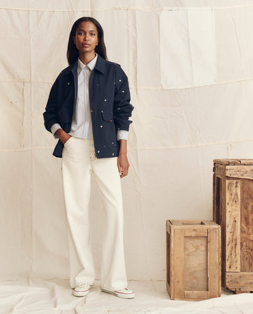 A woman stands indoors next to wooden crates, wearing THE GREAT. PS26’s The Folsom Jacket in navy with tossed floral embroidery, over a striped shirt and white wide-leg pants, against a neutral fabric backdrop.