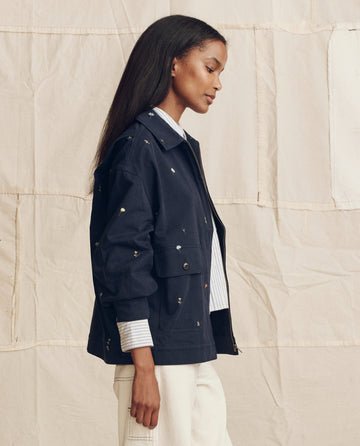 A woman stands in profile against a beige backdrop wearing THE GREAT. PS26 "The Folsom Jacket, Embroidered" in navy with tossed floral embroidery, styled with a striped shirt and white pants.