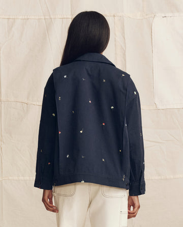 A person stands facing away, modeling THE GREAT. PS26's The Folsom Jacket. Embroidered in navy with tossed floral embroidery and off-white pants, against a light fabric backdrop.