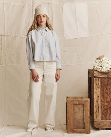 A woman in THE GREAT. PS26's The Mill Jean—Natural with Navy Stitching—striped shirt, and beanie stands indoors beside a crate and a box of white flowers, set against a neutral fabric backdrop.