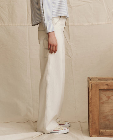 A person stands sideways next to a wooden box, wearing THE GREAT. PS26 The Mill Jean in Natural with Navy Stitching, a light blue striped shirt, and white sneakers against a neutral fabric backdrop.