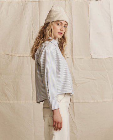 A woman with wavy blonde hair wears THE GREAT. PS26’s The Cropped Oxford Shirt in Soft Blue Stripe and a white knit beanie, paired with light-colored pants, standing in profile against a beige fabric backdrop.