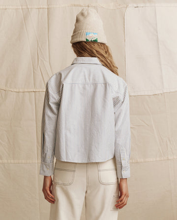 A person stands with their back to the camera, wearing THE GREAT. PS26 The Cropped Oxford Shirt in Soft Blue Stripe, a beige knit hat, and off-white pants against a neutral fabric backdrop.