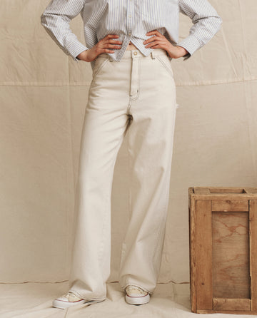 A person wears THE GREAT. PS26 The Mill Jean in natural with navy stitching, paired with a blue and white striped shirt and white shoes, standing by a wooden crate against a beige backdrop.