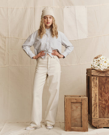 A woman stands indoors by wooden crates and white flowers, wearing THE GREAT. PS26's The Cropped Oxford Shirt in Soft Blue Stripe with white pants and a knit hat.