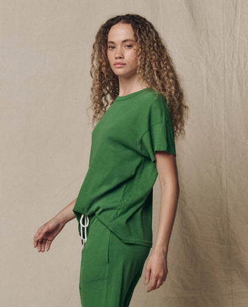 A person with long curly hair wears THE GREAT. SU26 SEASONAL CORE The Boxy Crew. Solid -- Fennel t-shirt and matching green pants, standing before a beige fabric backdrop.