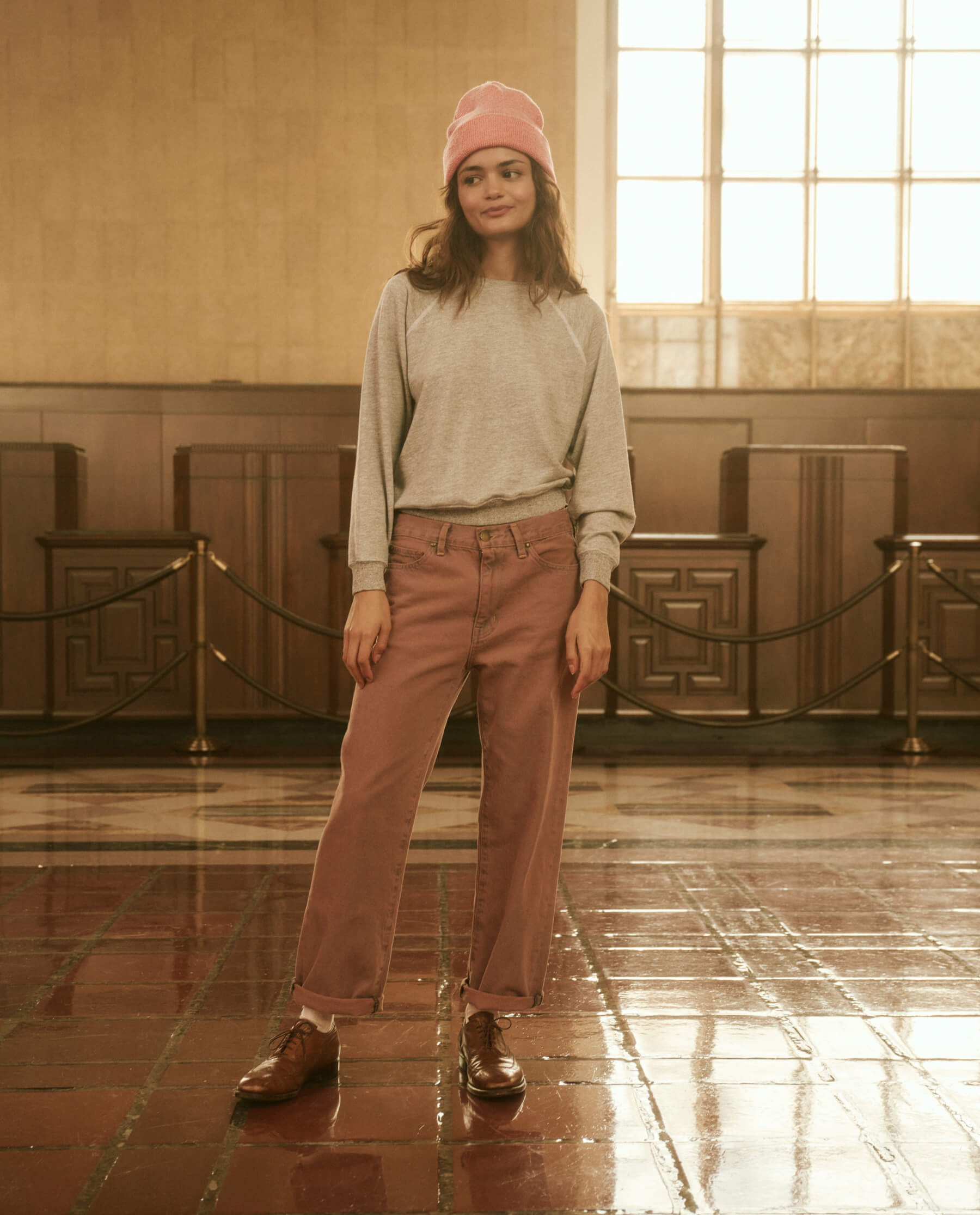 A person stands indoors on a glossy floor, wearing a gray sweatshirt, a pink beanie, The Billy Jean Clean Hem in Musky Pink by THE GREAT., and brown shoes with a relaxed silhouette. Their hands are in their pockets against a backdrop of wooden paneling and a large window.