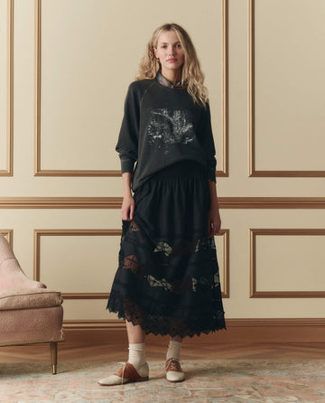 A woman stands indoors wearing THE GREAT. SP26 D2 The College Sweatshirt W/ Shadow Oak Graphic in washed black, a long black skirt with lace panels, beige socks, and two-tone oxford shoes.