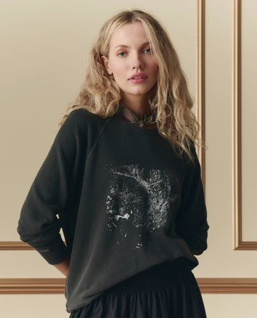 A woman with long blonde hair stands indoors wearing THE GREAT. SP26 D2 The College Sweatshirt W/ Shadow Oak Graphic in washed black and a patterned scarf around her neck.