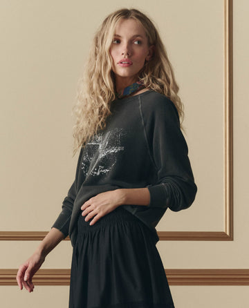 A woman with long, wavy blonde hair wears THE GREAT. SP26 D2 The College Sweatshirt W/ Shadow Oak Graphic in washed black and a black skirt, standing before a beige wall with decorative molding.