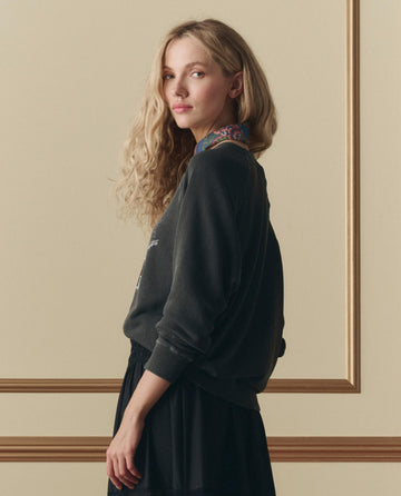 A woman with long, wavy blonde hair stands sideways against a beige wall, wearing THE GREAT. SP26 D2's The College Sweatshirt W/ Shadow Oak Graphic in washed black and a black skirt.