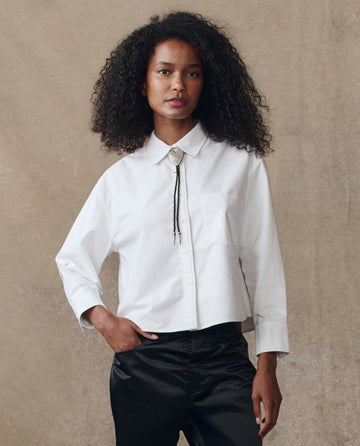 A person with curly hair wears THE GREAT. H25 COLLECTION The Cropped Oxford Shirt in white, styled with black pants and a bolo tie, standing against a plain beige background.