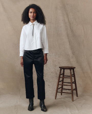A person stands indoors wearing THE GREAT. H25 COLLECTION's The Cropped Oxford Shirt in white, paired with black pants and boots, next to a wooden stool against a plain brown backdrop.