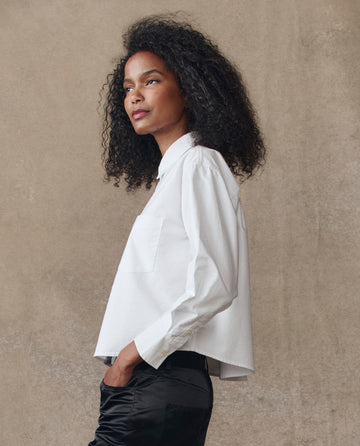 A person with curly hair models THE GREAT. H25 COLLECTION's The Cropped Oxford Shirt in white with tailored details and black pants, standing in profile against a plain beige background.