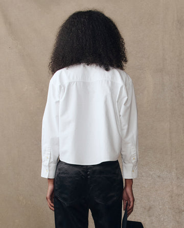 A person with curly hair wears THE GREAT. H25 COLLECTION The Cropped Oxford Shirt in white and black pants, standing with their back to the camera against a neutral background.