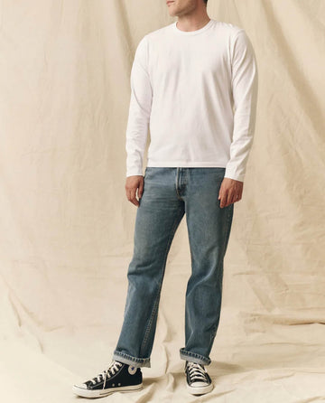 A person wearing THE GREAT. FALL 23 MEN The Men's Pure Knits Long Sleeve Slim Crew in True White, blue jeans, and black Converse sneakers stands against a beige fabric backdrop.