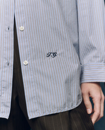 A person wearing THE GREAT. F25 COLLECTORS The Menswear Button Up in Alumni Stripe, a blue pinstriped shirt with "T.G." embroidery above the hem, paired with brown pinstriped pants.