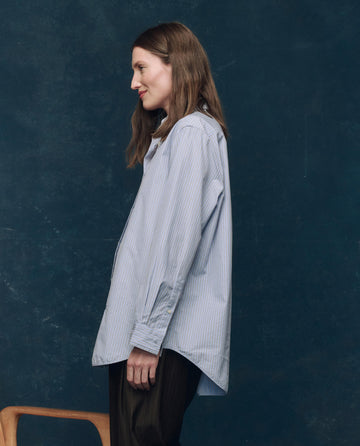 A woman stands in profile by a wooden chair, wearing THE GREAT. F25 COLLECTORS The Menswear Button Up. -- Alumni Stripe, a loose light blue striped vintage-style shirt, and dark pants, against a dark blue textured background.