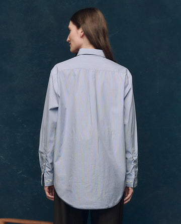 A person with long brown hair stands facing away, wearing THE GREAT. F25 COLLECTORS The Menswear Button Up -- Alumni Stripe and dark pants against a dark blue background.