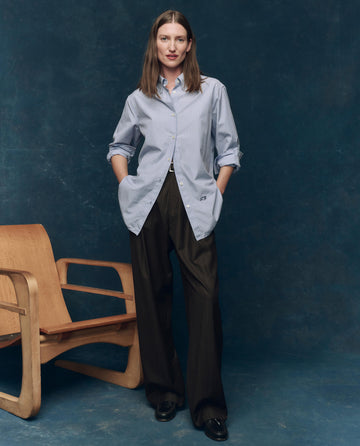 A person stands indoors in THE GREAT. F25 COLLECTORS The Menswear Button Up -- Alumni Stripe, styled with dark wide-leg trousers and black shoes, next to a wooden chair against a dark blue backdrop.