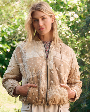 A woman stands outdoors with greenery behind her, wearing THE GREAT. SP26 TGO’s Reversible Ultra Light Puffer in Khaki Bandana w/ Cream, hands in her pockets.