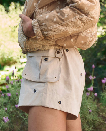 A person stands outdoors among green foliage and pink flowers, wearing THE GREAT. SP26 TGO's The Embroidered Cargo Short in cream with black daisy embroidery and a large pocket, paired with a light brown patterned jacket.