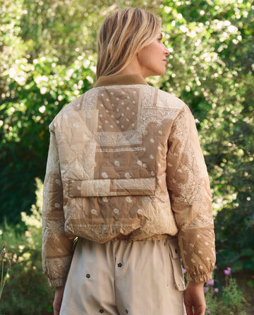 A person stands outdoors with green foliage in the background, wearing THE GREAT. SP26 TGO's The Reversible Ultra Light Puffer in Khaki Bandana W/ Cream and beige shorts with black prints.