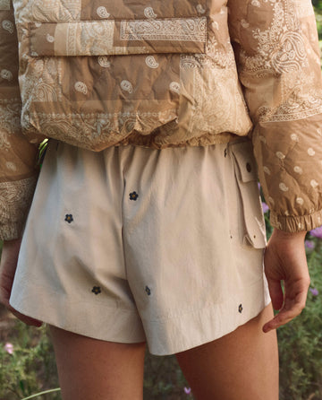 A person stands outdoors in THE GREAT. SP26 TGO The Embroidered Cargo Short in Cream with Black Daisies, paired with a patterned beige jacket, surrounded by greenery.