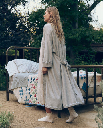 Wearing THE GREAT. CORE SLEEP's The Sweatshirt Robe in Light Heather Grey, a person stands by an outdoor bed with a patchwork quilt, surrounded by trees and greenery.