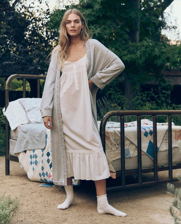 A woman in a long white nightgown and THE GREAT. CORE SLEEP’s The Sweatshirt Robe in Light Heather Grey stands outdoors beside a vintage metal bed topped with patterned quilts.